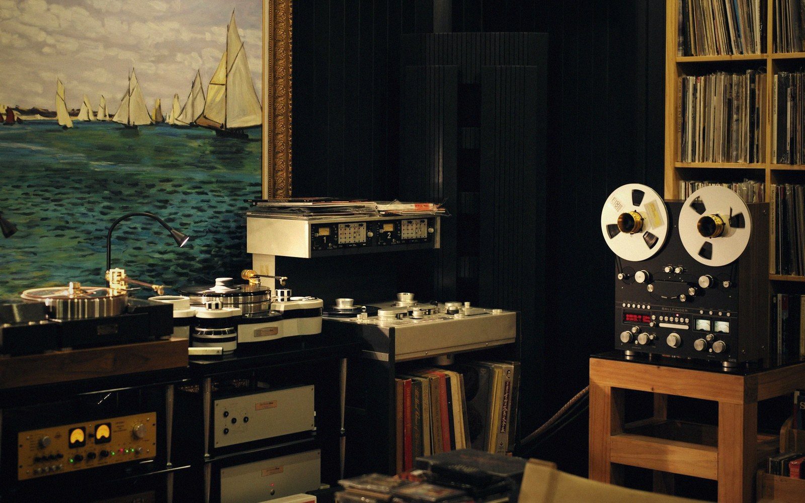 Reel-to-reel tape player and turntables in a listening room with vinyl shelves and a sailboat painting