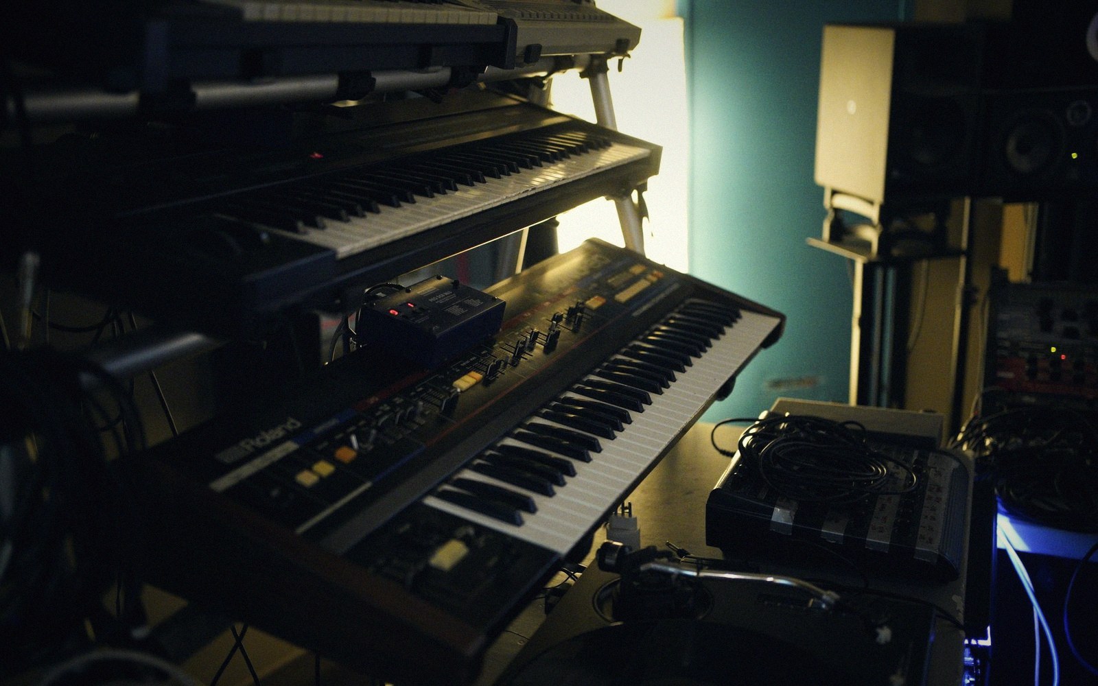 Stacked synthesizer keyboards and drum machine in a dimly lit studio