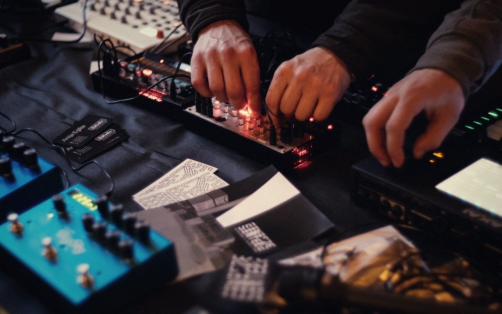 Hands adjusting knobs on a modular synthesizer surrounded by pedals and printed flyers