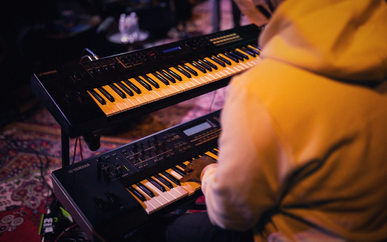 A hooded musician plays two stacked Yamaha keyboards during a dimly lit performance
